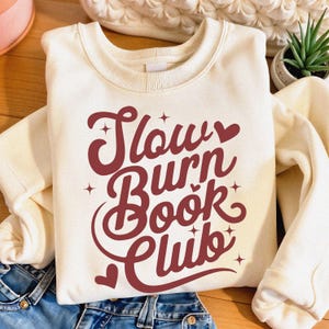 Può includere: Felpa color crema con la scritta "Slow Burn Book Club" in corsivo bordeaux. Il design include cuori e stelle. La felpa è piegata sopra dei jeans blu.