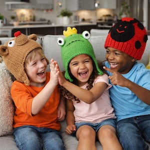Pode incluir: Três crianças usando chapéus de crochê feitos à mão, cada um com um design de animal único. Os chapéus incluem uma rena marrom, um sapo verde com uma coroa amarela e uma joaninha vermelha. As crianças estão rindo e sentadas em um sofá cinza.