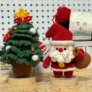 May include: Crocheted Christmas decorations featuring a green tree with a yellow star and a Santa Claus figure in red and white. The tree has white ball ornaments and gold bells. Santa holds a brown sack.
