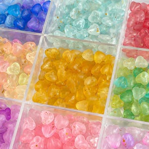 May include: A clear plastic storage container with nine square compartments filled with small, heart-shaped beads in various translucent colors including blue, mint green, red, peach, yellow, lime green, purple, pink, and lavender.