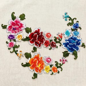 May include: A floral embroidery patch featuring a vibrant arrangement of multi-coloured peonies and smaller blossoms in shades of pink, red, orange, blue, and yellow, connected by intricate green leafy stems and vines on a light fabric background.