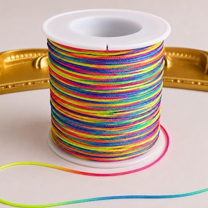 Può includere: Una bobina di cordino multicolore con un vivace motivo arcobaleno di fili giallo neon, rosa brillante, blu elettrico e arancione avvolti strettamente attorno a un cilindro di plastica bianca, con un singolo filo sciolto alla base.
