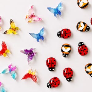 Op de afbeelding: Een verzameling kleine decoratieve bedels met doorschijnende, veelkleurige vlinders en ondoorzichtige, glanzende lieveheersbeestjes en bijen. De vlinders vertonen levendige gradiënten van roze, geel, blauw en paars, terwijl de insecten rood zijn met zwarte stippen of geel-zwart gestreepte patronen hebben.