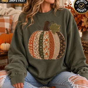 May include: An olive green crewneck sweatshirt featuring a large, central pumpkin graphic. The pumpkin is composed of vertical fabric panels with various autumn-themed patterns, including orange gingham, floral prints, and small pumpkin motifs.