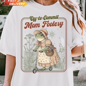May include: White t-shirt with a whimsical illustration. The design features a frog in a bonnet and dress riding a scooter, with the text "Off-to-Commit Mom Foolery" in a vintage style. The image has a retro, storybook aesthetic.