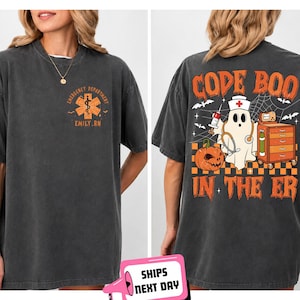 May include: A charcoal gray t-shirt featuring a small orange medical star logo on the front left chest. The back displays a large graphic of a ghost nurse, a jack-o'-lantern, and medical equipment with the text CODE BOO IN THE ER.