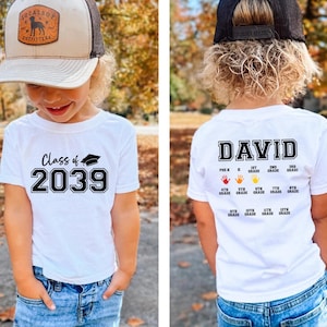 May include: White t-shirt with the text "Class of 2039" and a graduation cap graphic. The back of the shirt has the name "DAVID" and a series of handprints with grade levels. The child is wearing blue jeans and a baseball cap.