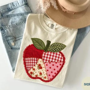 May include: An ivory t-shirt featuring a large apple-shaped applique made of various red and green fabric patches, including gingham, polka dot, and floral patterns. The shirt is paired with light blue denim jeans and a tan wide-brimmed hat.