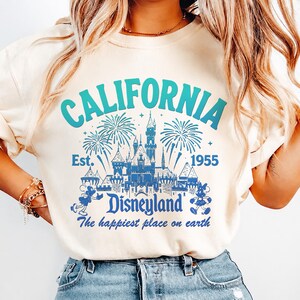May include: Cream-colored sweatshirt with a blue and teal graphic that reads "CALIFORNIA Disneyland" with a castle, fireworks, and Mickey Mouse. The text "Est. 1955" and "The happiest place on earth" are also included.