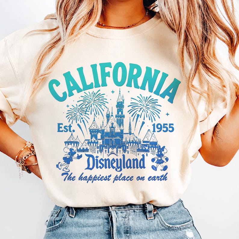 May include: Cream-colored sweatshirt with a blue and teal graphic that reads "CALIFORNIA Disneyland" with a castle, fireworks, and Mickey Mouse. The text "Est. 1955" and "The happiest place on earth" are also included.