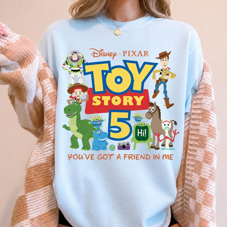 May include: A light blue crewneck sweatshirt featuring a colorful Disney Pixar Toy Story 5 graphic. The design includes characters like Buzz Lightyear, Woody, Jessie, Rex, Bullseye, and Forky, with the text "TOY STORY 5" and "YOU'VE GOT A FRIEND IN ME."
