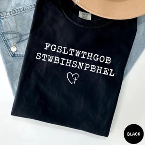 May include: Black t-shirt with white text and a heart with a cross symbol. The shirt is made of a soft material and is labeled "Comfort Colors". The shirt is paired with blue jeans and a tan hat.