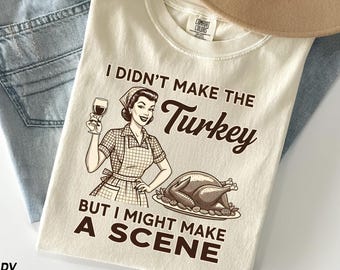 May include: Cream-colored t-shirt with the text "I Didn't Make The Turkey But I Might Make A Scene." The graphic features a vintage illustration of a woman holding a wine glass next to a roasted turkey.