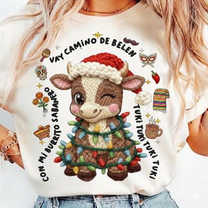 May include: White t-shirt featuring a cartoon cow wearing a Santa hat and wrapped in Christmas lights. The shirt includes text in Spanish, along with festive illustrations like a skull, maracas, and a coffee cup.