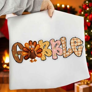 Könnte beinhalten: Ein weißes Kissen mit dem Wort "GOBBLE" in bunten, gemusterten Buchstaben. Neben dem Buchstaben "G" befindet sich ein Zeichentricktruthahn mit braunen, orangefarbenen und gelben Federn. Das Kissen wird von einer Person gehalten.