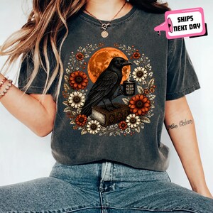 May include: A charcoal gray t-shirt featuring a graphic of a black crow perched on a book, holding a mug with the text IS HE DEAD YET? The crow is surrounded by a wreath of orange and white flowers against a large, round orange moon background.