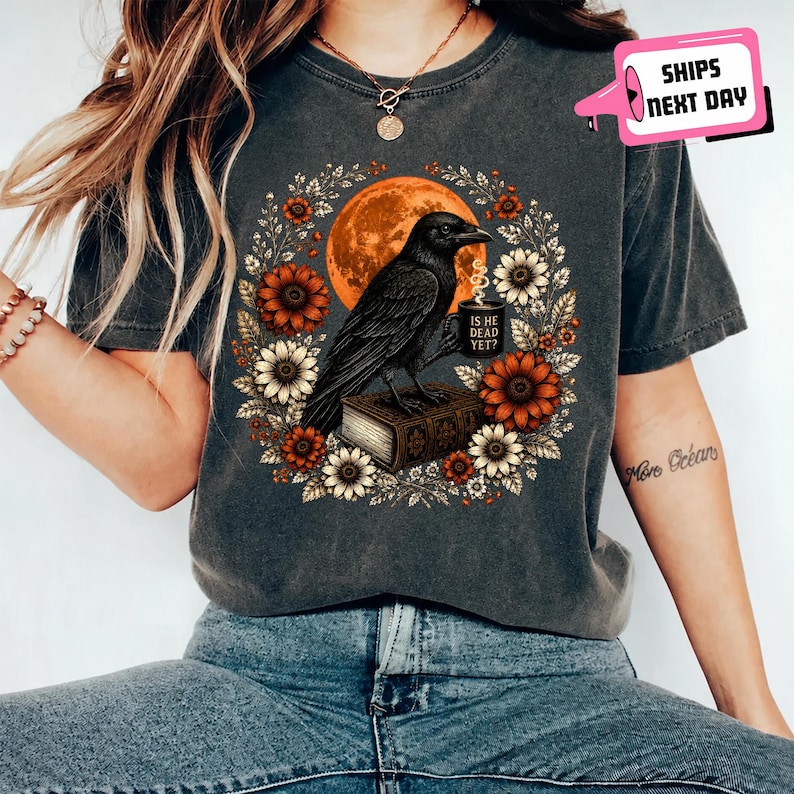 May include: A charcoal gray t-shirt featuring a graphic of a black crow perched on a book, holding a mug with the text IS HE DEAD YET? The crow is surrounded by a wreath of orange and white flowers against a large, round orange moon background.