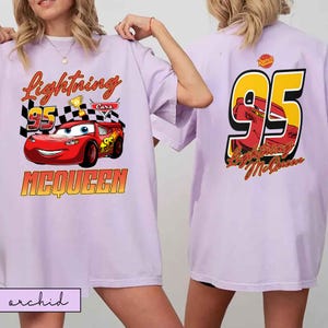 Może przedstawiać: Jasnofioletowa koszulka z Lightning McQueen z filmu Auta. Prz&oacute;d przedstawia napis "Lightning 95 McQueen" z motywem samochodu wyścigowego. Tył koszulki pokazuje duży "95" z wizerunkiem McQueena.