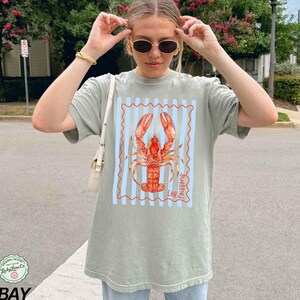 以下が含まれることがあります： セージグリーンのTシャツ。赤色のロブスターのイラストが、青と白のストライプのフレーム内に描かれています。ロブスターのデザインには「Les Poissons」の文字が含まれています。Tシャツは半袖で、ゆったりとしたフィット感です。