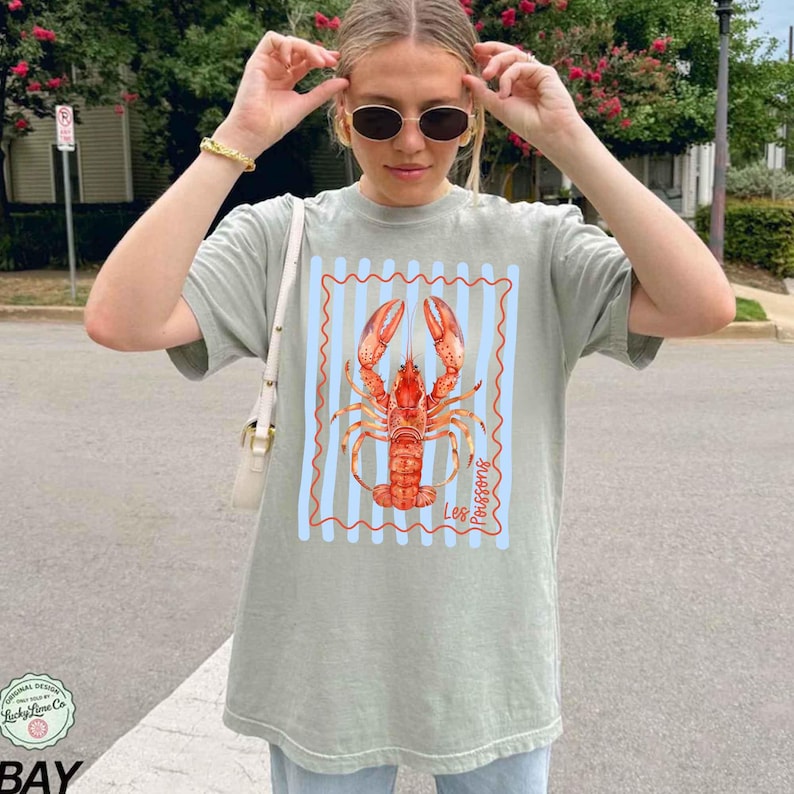 以下が含まれることがあります： セージグリーンのTシャツ。赤色のロブスターのイラストが、青と白のストライプのフレーム内に描かれています。ロブスターのデザインには「Les Poissons」の文字が含まれています。Tシャツは半袖で、ゆったりとしたフィット感です。