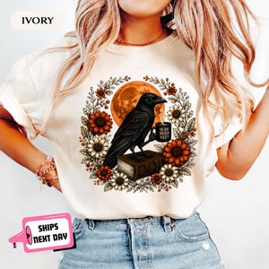 May include: An ivory t-shirt featuring a graphic of a black raven perched on a book, holding a mug with the text 'IS HE DEAD YET?'. The raven is surrounded by a circular wreath of sunflowers and autumn leaves against a large orange moon.
