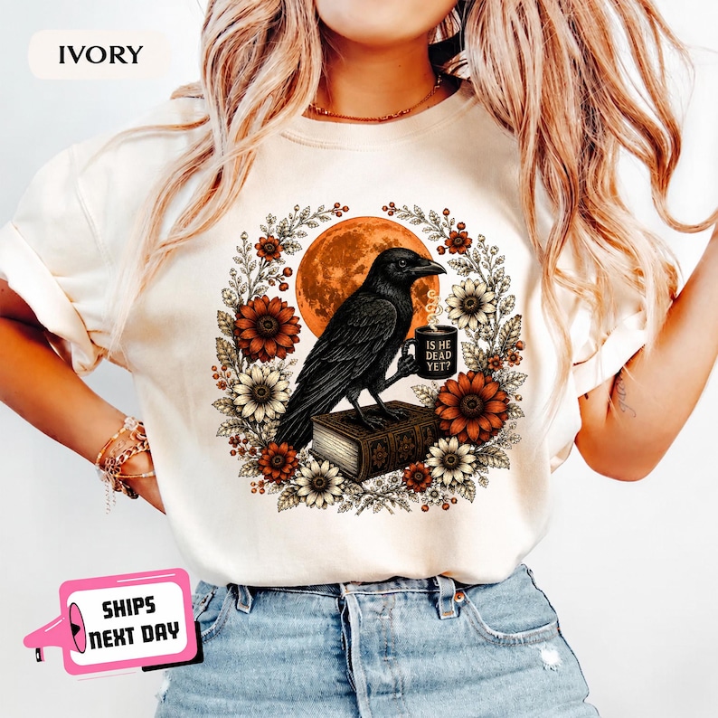 May include: An ivory t-shirt featuring a graphic of a black raven perched on a book, holding a mug with the text 'IS HE DEAD YET?'. The raven is surrounded by a circular wreath of sunflowers and autumn leaves against a large orange moon.