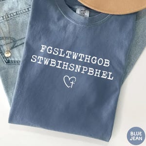 May include: A blue jean Comfort Colors t-shirt with white text and a heart-shaped cross graphic. The shirt's text is an encrypted message. A tan hat and a silver necklace are visible in the background.