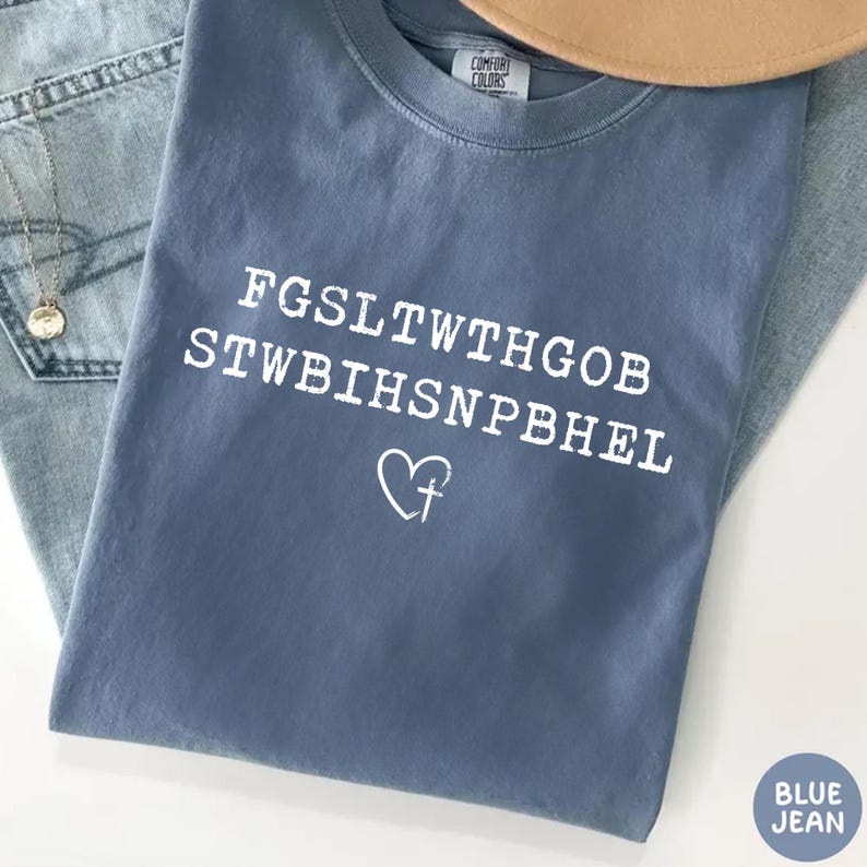 May include: A blue jean Comfort Colors t-shirt with white text and a heart-shaped cross graphic. The shirt's text is an encrypted message. A tan hat and a silver necklace are visible in the background.