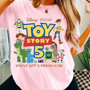 May include: A light pink t-shirt featuring a colorful graphic of Disney Pixar Toy Story characters including Buzz Lightyear, Woody, Jessie, Rex, and Forky. The design includes the text Toy Story 5 and the phrase You've Got A Friend In Me.