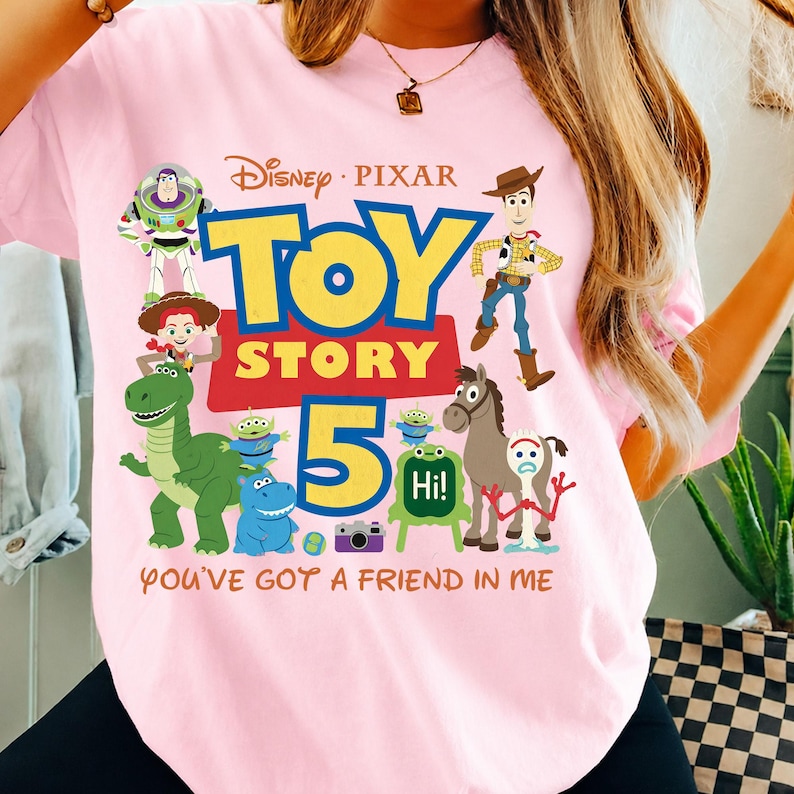 May include: A light pink t-shirt featuring a colorful graphic of Disney Pixar Toy Story characters including Buzz Lightyear, Woody, Jessie, Rex, and Forky. The design includes the text Toy Story 5 and the phrase You've Got A Friend In Me.