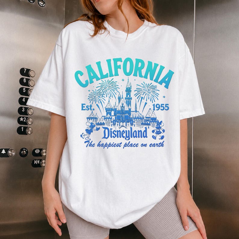 May include: White t-shirt with a blue and teal graphic that says "CALIFORNIA Disneyland" with fireworks and a castle design. The text "Est. 1955" and "The happiest place on earth" are also included.