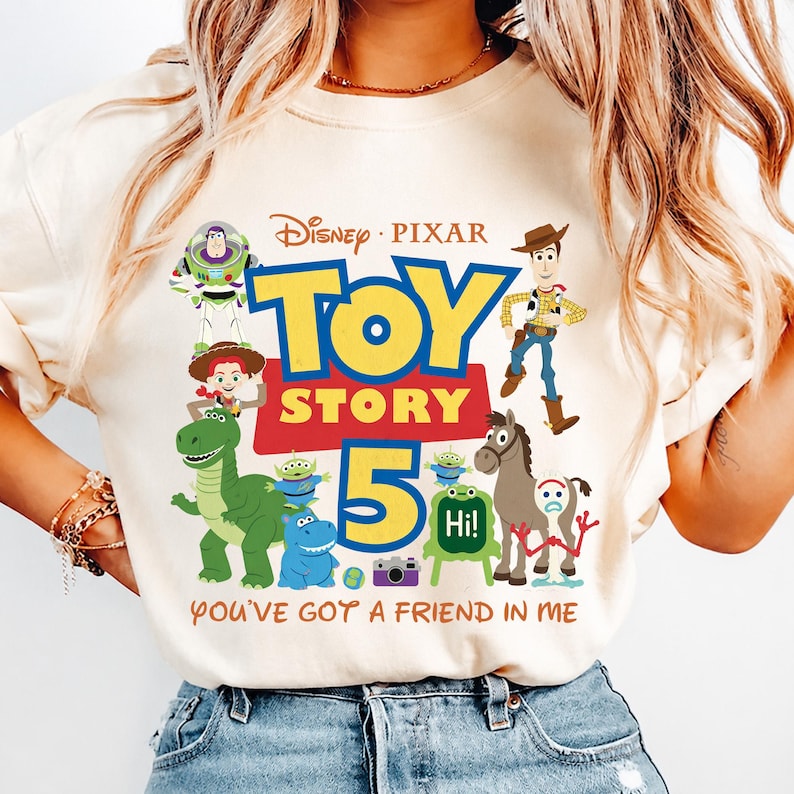 May include: A cream-colored t-shirt featuring a Disney Pixar Toy Story 5 graphic. The design includes characters like Woody, Buzz Lightyear, Jessie, Rex, and Forky, with the text Toy Story 5 and You've Got A Friend In Me printed in colorful fonts.