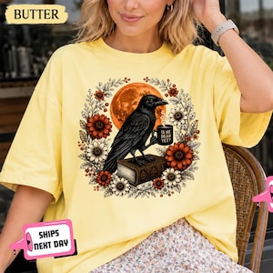 May include: A butter yellow short-sleeve t-shirt featuring a graphic of a black crow perched on a book, holding a mug that reads IS DEAD YET? The crow is surrounded by a circular wreath of orange and white flowers against a large orange moon.