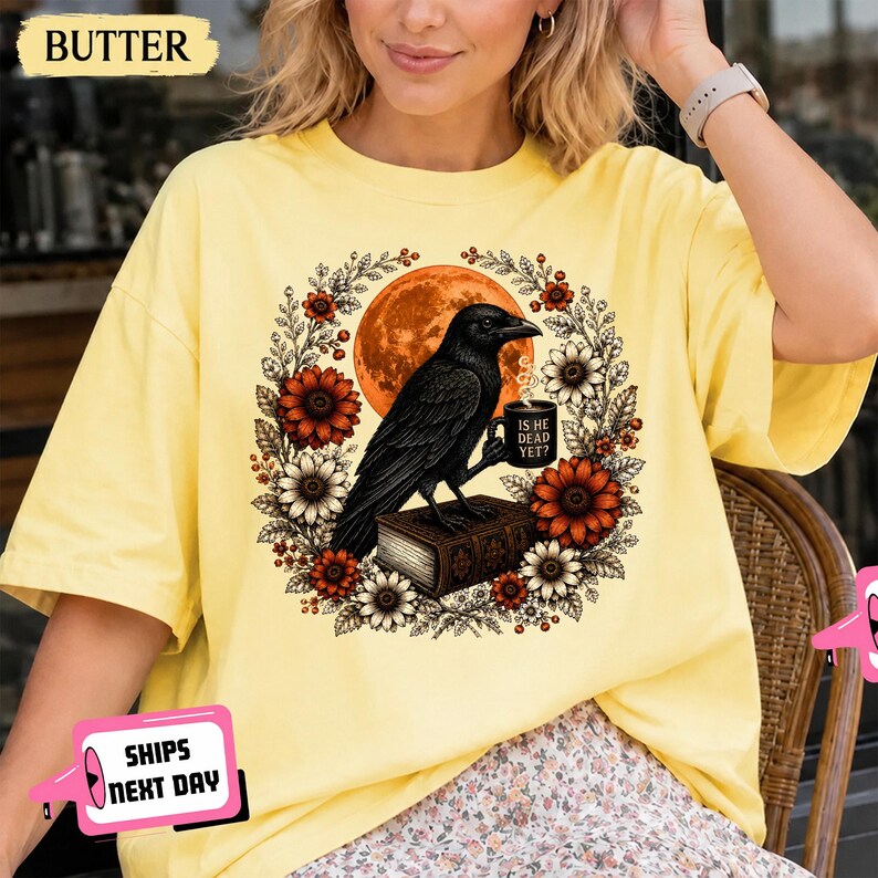 May include: A butter yellow short-sleeve t-shirt featuring a graphic of a black crow perched on a book, holding a mug that reads IS DEAD YET? The crow is surrounded by a circular wreath of orange and white flowers against a large orange moon.