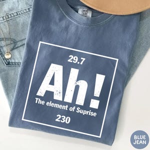 May include: Blue jean colored t-shirt with a white graphic. The graphic resembles a periodic table element with the text "Ah! The element of Suprise 230" in a white box. The shirt is folded on top of blue jeans.