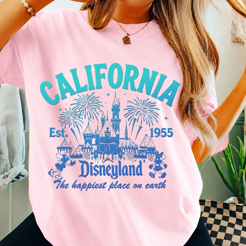 May include: Pink t-shirt with a blue graphic that says "CALIFORNIA Disneyland The happiest place on earth Est. 1955". The graphic features a castle, fireworks, and Mickey Mouse.