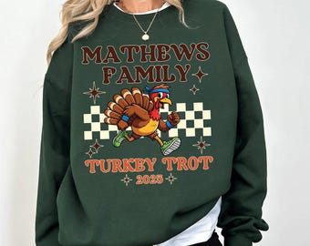 May include: Dark green sweatshirt with the text "MATHEWS FAMILY" and "TURKEY TROT 2025". The graphic features a cartoon turkey running with a checkered background and starburst accents. The sweatshirt is a long-sleeved crew neck.