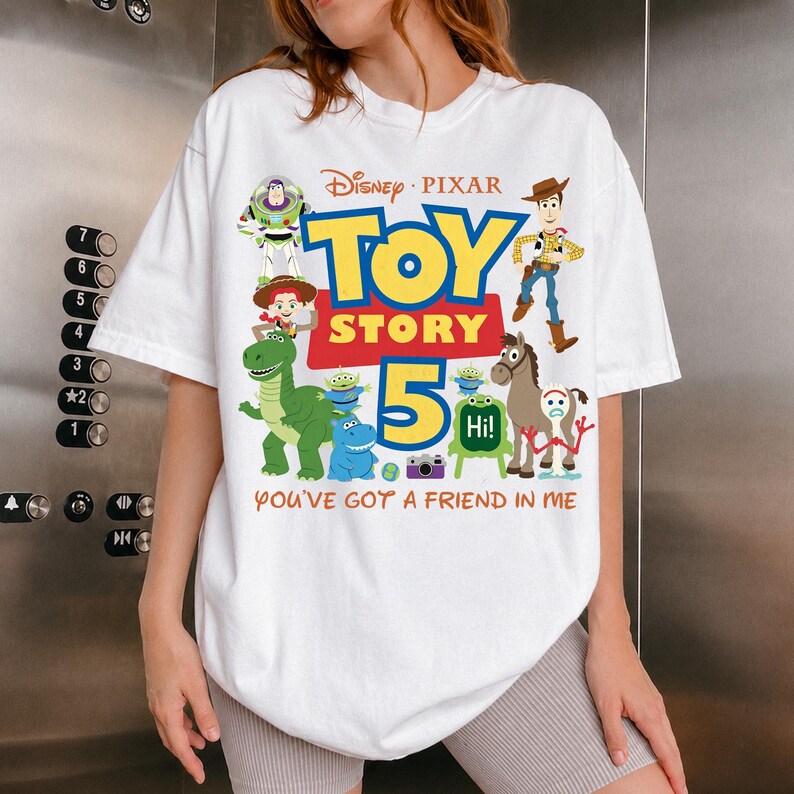 May include: A white short-sleeved t-shirt featuring a colorful graphic print of Disney Pixar Toy Story 5 characters including Woody, Buzz Lightyear, Jessie, Rex, Bullseye, and Forky, with the text Toy Story 5 and You've Got A Friend In Me.