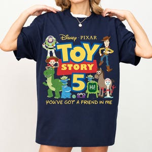 May include: A navy blue short-sleeve t-shirt featuring a colorful Disney Pixar Toy Story 5 graphic. The design includes characters like Woody, Buzz Lightyear, Rex, and Forky, with the text "TOY STORY 5" and "YOU'VE GOT A FRIEND IN ME" printed in yellow.