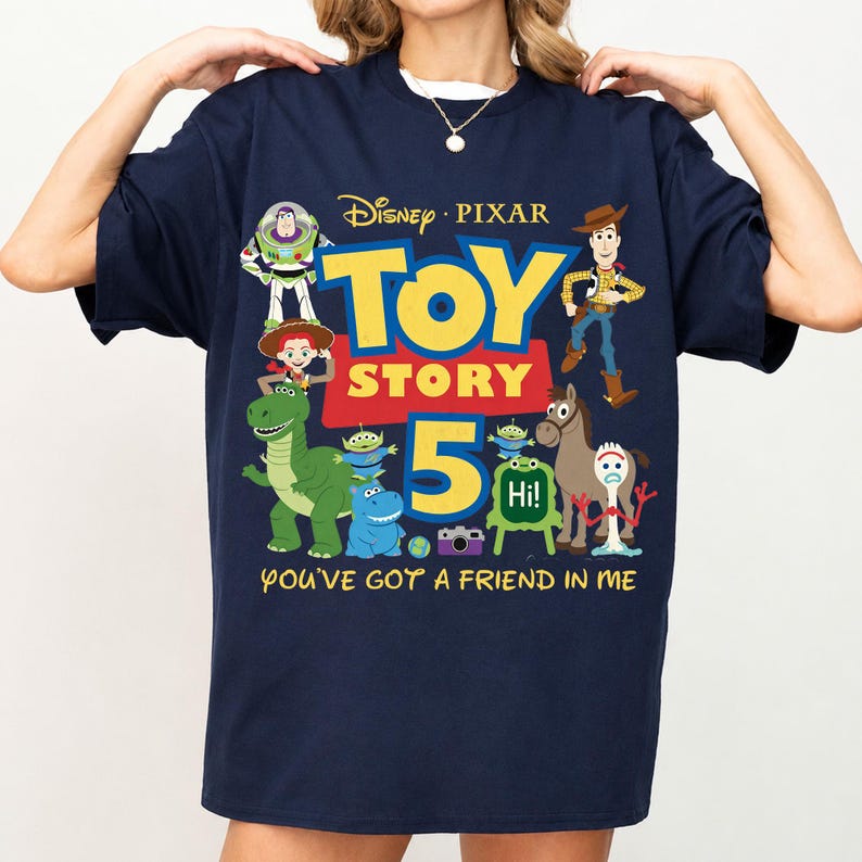 May include: A navy blue short-sleeve t-shirt featuring a colorful Disney Pixar Toy Story 5 graphic. The design includes characters like Woody, Buzz Lightyear, Rex, and Forky, with the text "TOY STORY 5" and "YOU'VE GOT A FRIEND IN ME" printed in yellow.