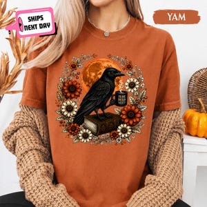 May include: A rust-colored short-sleeve t-shirt featuring a graphic of a black crow perched on a book, holding a mug with the text 'IS HE DEAD YET?'. The crow is surrounded by a circular wreath of orange and white sunflowers and a large orange moon.