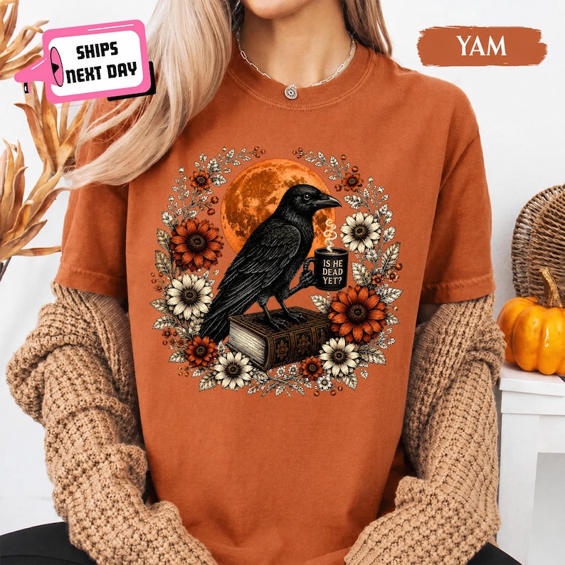 May include: A rust-colored short-sleeve t-shirt featuring a graphic of a black crow perched on a book, holding a mug with the text 'IS HE DEAD YET?'. The crow is surrounded by a circular wreath of orange and white sunflowers and a large orange moon.
