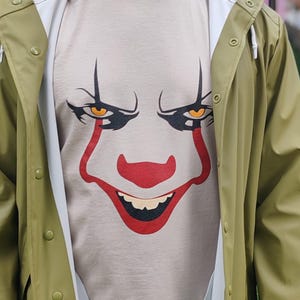 Può includere: Una maglietta beige con una grafica del volto di un clown in rosso e nero. La persona indossa un impermeabile verde con fodera bianca. L'impermeabile ha una chiusura a bottoni.