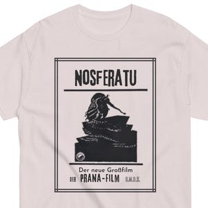 Pode incluir: Uma t-shirt de manga curta rosa claro com uma impressão gráfica preta do cartaz do filme Nosferatu. O design inclui o texto NOSFERATU, Der neue Großfilm e DER PRANA-FILM G.M.B.H. dentro de uma borda retangular na frente da peça.