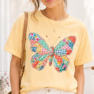 以下が含まれることがあります： カラフルなパッチワークの蝶のデザインが施された黄色のTシャツ。白いショートパンツと合わせています。晴れた日に屋外で撮影された画像です。