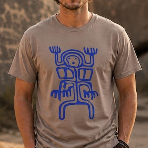 Op de afbeelding: Een T-shirt met korte mouwen en een ronde hals in een lichtbeige kleur, voorzien van een opvallende, felblauwe grafische print op de borst. De afbeelding toont een gestileerd, symmetrisch figuur met opgeheven armen en benen.