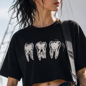 Op de afbeelding: Zwart, gecropt T-shirt met een afbeelding van drie tanden. Het shirt wordt gecombineerd met een zwarte jeans en een gitaar. De foto is buiten genomen met een heldere achtergrond.
