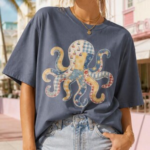 Peut inclure: Un t-shirt bleu marine avec un motif de pieuvre colorée. La pieuvre a des tentacules de style patchwork avec des motifs nautiques. Le t-shirt est associé à un short en jean. L'arrière-plan comprend des palmiers et un bâtiment.