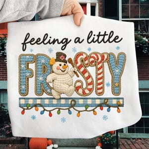 K&ouml;nnte beinhalten: Wei&szlig;er Stoff mit dem Text "feeling a little FROSTY" in schwarzer Schrift. Das Wort "FROSTY" ist in einem blau-roten Design mit einem Schneemann und einer Zuckerstange gehalten. Ein blau-wei&szlig; karierter Rand mit bunten Lichtern und Schneeflocken vervollst&auml;ndigt das Design.