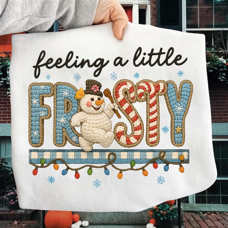 K&ouml;nnte beinhalten: Wei&szlig;er Stoff mit dem Text "feeling a little FROSTY" in schwarzer Schrift. Das Wort "FROSTY" ist in einem blau-roten Design mit einem Schneemann und einer Zuckerstange gehalten. Ein blau-wei&szlig; karierter Rand mit bunten Lichtern und Schneeflocken vervollst&auml;ndigt das Design.